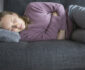 Sick woman lying on grey sofa at home, having legs bent